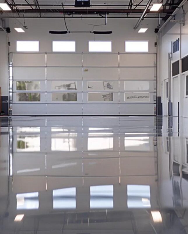 Epoxy flooring in a spacious garage with a glossy finish, showcasing reflections and bright overhead lighting, emphasizing high-quality flooring solutions by Pristine Coatings.